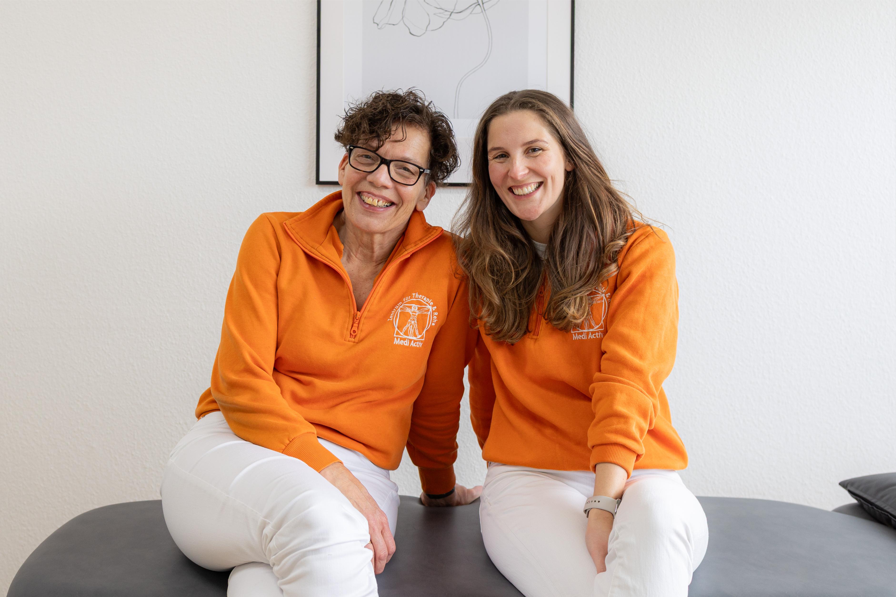 Zwei Frauen sitzen nebeneinander auf einer schwarzen Liege und tragen orangefarbene Pullover mit weißer Aufschrift und weiße Hosen