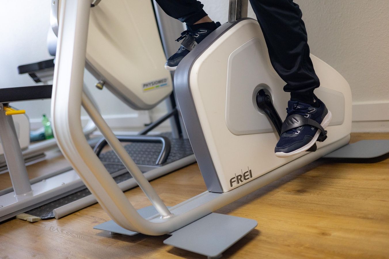 Beine einer Person mit Sportschuhen auf einem stationären Fahrrad im Fitnessraum