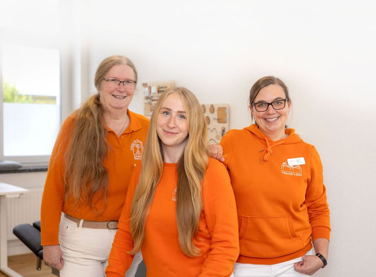 Drei Frauen in orangefarbenen Oberteilen stehen in einem hellen Raum mit Fenster und Möbeln im Hintergrund