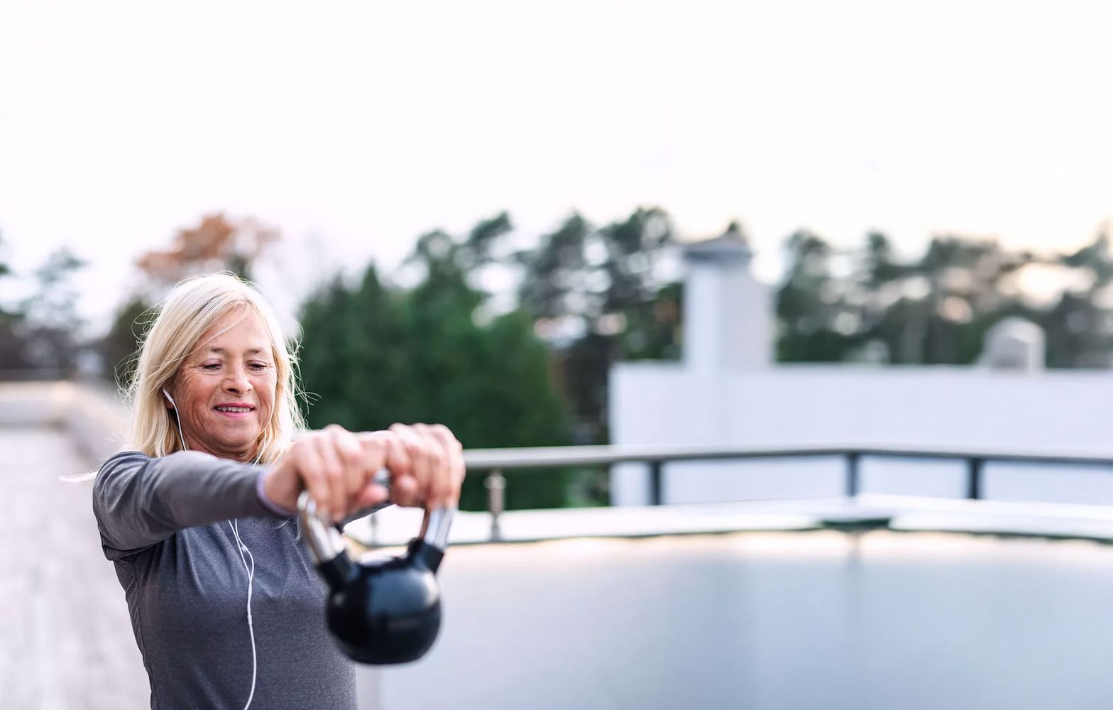 Frau in grauem Sportshirt hält mit ausgestreckten Armen eine Kettlebell vor sich