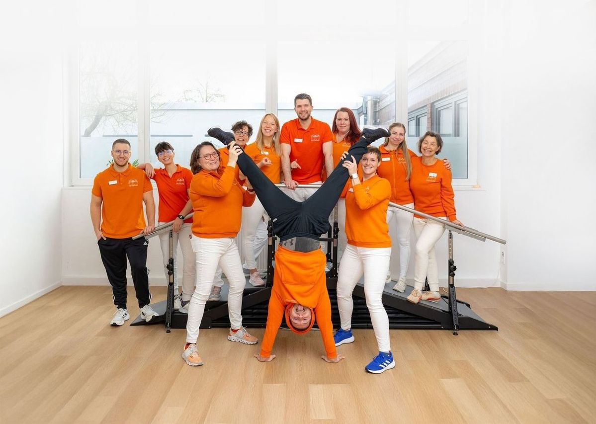 Gruppe von neun Personen in orangefarbenen Oberteilen in einem hellen Raum, eine Person macht einen Handstand, andere halten sie an den Beinen, im Hintergrund sind zwei parallele Barren zu sehen.