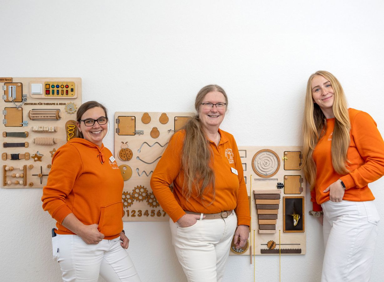 Drei Personen in orangen Pullovern und weißen Hosen stehen vor einer Wand mit verschiedenen hölzernen Lern- und Spieltafeln